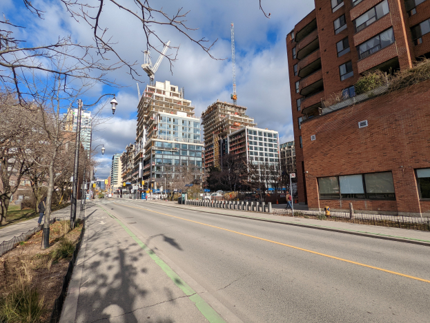Eine Stadtstraße mit hohen Gebäuden, Straßenlaternen, Strommasten, Stromkabeln, Kraftfahrzeugen, Fußgängern, Geländern, Bäumen, Pflanzen und einem bewölkten Himmel.