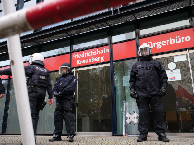 Eine Gruppe von Polizisten in Helmen und Handschuhen steht vor einem Gebäude mit Glaswänden und -türen, mit einem Pfahl links und Schildern im Hintergrund, die "Friedrichshain Kreuzberg" und Papiere an den Glaswänden angeklebt zeigen.