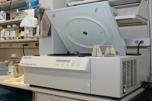 Ein Laborarbeitsplatz mit einem Mikrotom-Gerät oben drauf, Flaschen und andere Gegenstände auf Gestellen links daneben und eine Wand im Hintergrund.