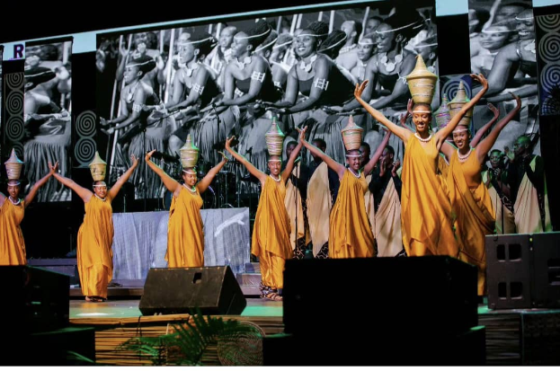 Eine Gruppe von Menschen in gelben Kleidern steht auf einer Bühne mit Lautsprechern auf beiden Seiten, die verschiedene Gegenstände halten, mit einem großen Bildschirm im Hintergrund und einer Pflanze am unteren Bildrand.