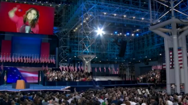 Eine große Menschenmenge sitzt vor einer Bühne mit einem Podium, einem Mikrofon, Flaggen, einem Bildschirm mit einem Bild einer Frau und Beleuchtungsausrüstung, vermutlich für eine Präsidentenrede.