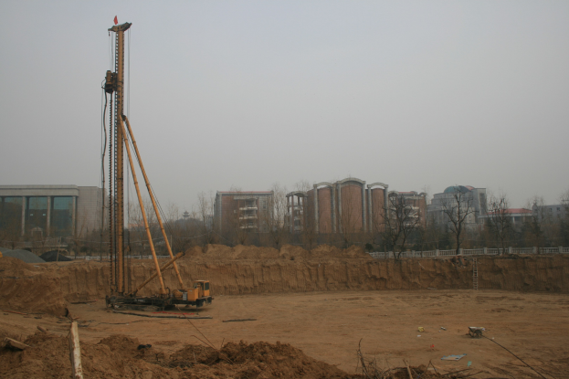 Eine Baustelle mit einem Kran in der Mitte, umgeben von Gebäuden, Bäumen und einem klaren blauen Himmel, mit Erdreich bedecktem Boden und verstreuten Gegenständen.