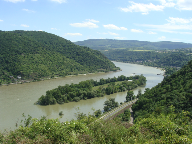 Panoramablick auf den sich durch eine grüne Landschaft schlängelnden Rhein mit einer Straße daneben, unter einem teilweise bewölkten Himmel.