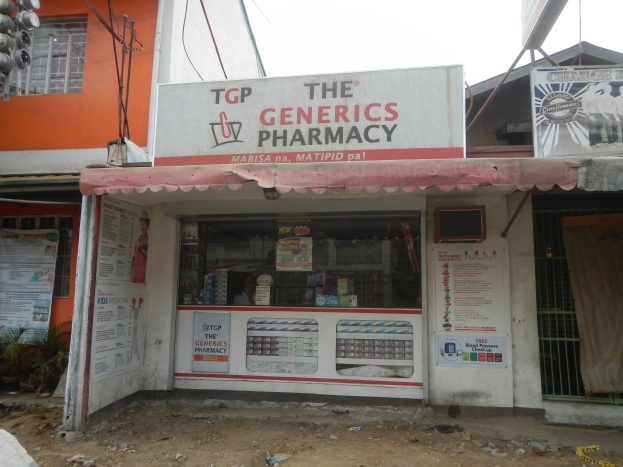 Ein Apotheken-Schild mit der Aufschrift "The Generics Pharmacy" vor einem Gebäude, umgeben von städtischen Elementen unter einem klaren blauen Himmel.