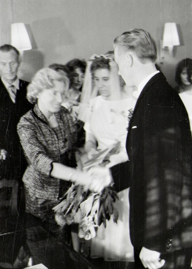 Ein Schwarz-Weiß-Foto eines Brautpaares beim Austausch der Eheringe, mit der Braut in einem weißen Kleid und Schleier, die einen Strauß hält, und dem Bräutigam im Anzug und Krawatte, mit Wandlampen im Hintergrund.