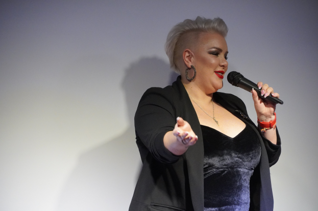 Eine Frau in einem schwarzen Kleid und Blazer steht selbstbewusst auf einer Bühne, hält ein Mikrofon und hat einen entschlossenen Gesichtsausdruck und eine starke Körperhaltung.