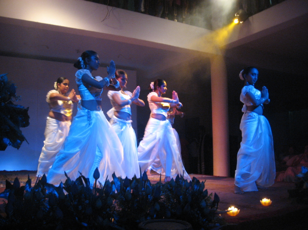 Eine Gruppe von Menschen, die auf einer Bühne mit Pflanzen davor und einer Gruppe von Menschen, die zuschauen, tanzen.