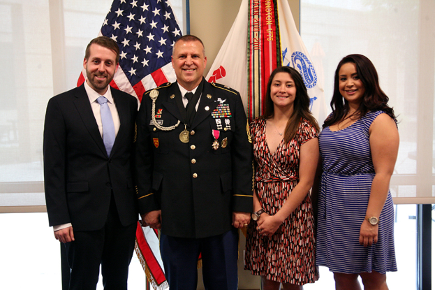 Zwei Frauen in Kleidern, eine Person in Uniform und ein Mann in Anzug stehen und lächeln drinnen mit Jalousien und Fahnen im Hintergrund.