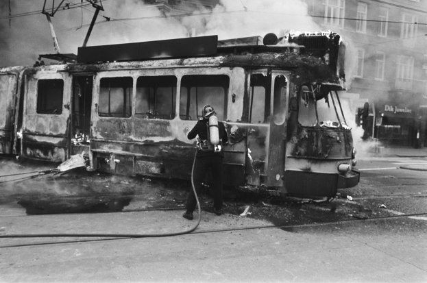 Schwarzes und wei├čes Bild eines Mannes in einem Helm, der eine Pfeife h├Ąlt, neben einer ausgebrannten Stra├čenbahn auf einer Stra├če, mit aufsteigendem Rauch und einem Gebäude im Hintergrund.