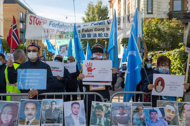 Eine Gruppe von Menschen mit Masken und Schildern und Plakaten steht vor einem Zaun mit Fotos und Text, protestiert in Istanbul, Türkei, mit Gebäuden, Bäumen und einem Laternenpfahl im Hintergrund.