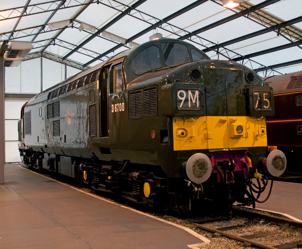 Ein Zug fährt auf den Schienen neben einem Bahnsteig mit einem Mast, der Metallstangen mit Lampen trägt.