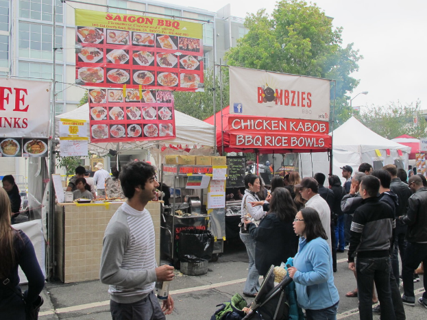 Menschen stehen auf einer Straße außerhalb der Stadt, mit einem Mann in einem grauen und weißen T-Shirt, der eine Wasserflasche hält, und einer Frau in einer blauen Jacke, die einen Kinderwagen schiebt. Im Hintergrund sind Stände mit weißen und roten Zelten und bunten Bannern sowie Gebäude, Bäume und eine Straßenlaterne zu sehen.
