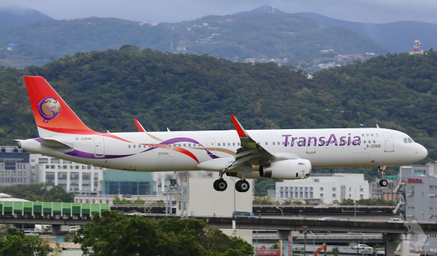 Ein Airbus A320-200 der TransAsia von Hong Kong International Airport mit einer Brücke im Vordergrund, Fahrzeugen auf der Straße darunter und Gebäuden, Bäumen und Bergen im Hintergrund unter einem bewölkten Himmel.