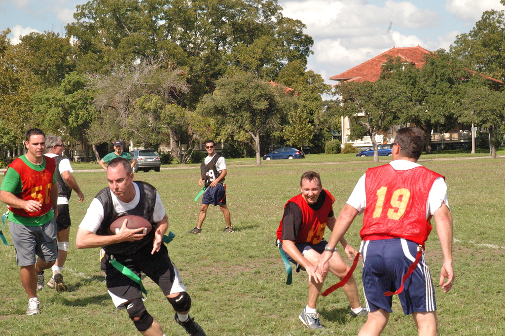 Menschen spielen American Football auf einem grünen Feld mit Bäumen, einem Haus und Autos im Hintergrund.