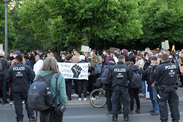 Große Gruppe von Menschen auf einer Straße, einige halten Protestschilder, andere tragen Mützen und Taschen, mit einem Fahrrad im Vordergrund und Bäumen und einem Pfahl im Hintergrund.
