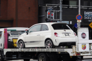 Ein weißes Auto wird von einem Abschleppwagen auf einer Straße mit Gebäuden, Mästen, Verkehrszeichen und Schildern im Hintergrund abgeschleppt.