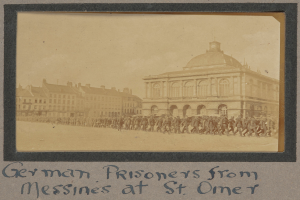 Deutsche Kriegsgefangene aus Messines vor einem Gebäude mit einem klaren blauen Himmel im Hintergrund, unten ist Text sichtbar.