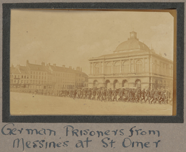 Deutsche Kriegsgefangene aus Messines vor einem Gebäude mit einem klaren blauen Himmel im Hintergrund, unten ist Text sichtbar.