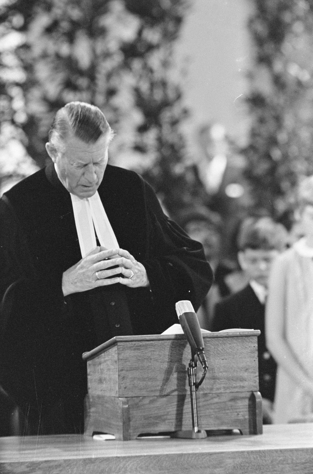 Ein Mann in einem schwarzen und weißen Gewand steht an einem Rednerpult mit einem Mikrofon und spricht zu einer kleinen Gruppe von Menschen mit Bäumen im Hintergrund.