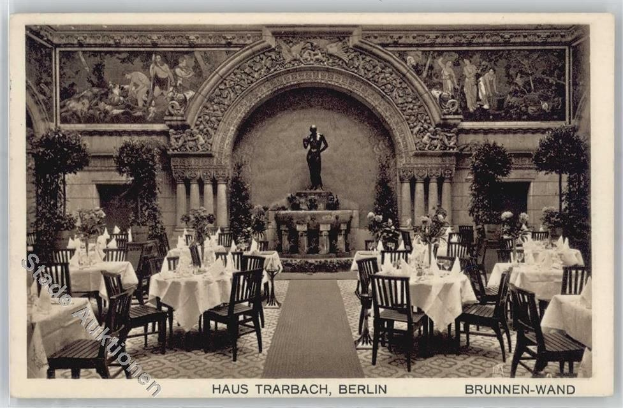 Schwarzes und weißes Foto eines alten Berliner Restaurantinnern mit Tischen, die mit Vasen, einer Statue im Hintergrund, Wandskulpturen, Pflanzen und Text am Boden dekoriert sind.