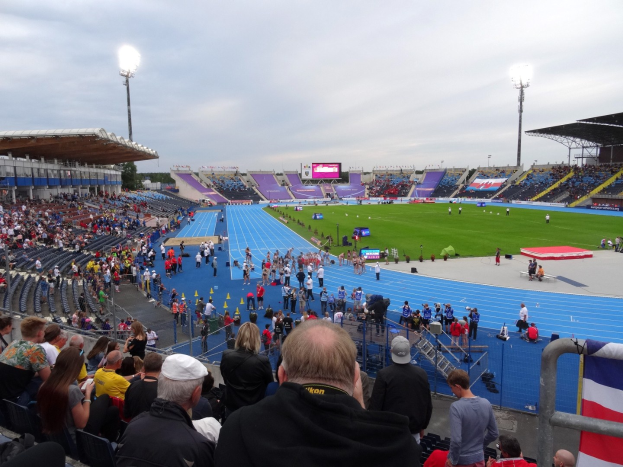 Großes Stadion voller Zuschauer bei einem Fußballspiel, mit sitzenden und stehenden Zuschauern, Geländern, einer Fahne, Flutlicht, einem Bildschirm, Bäumen und einem bewölkten Himmel.
