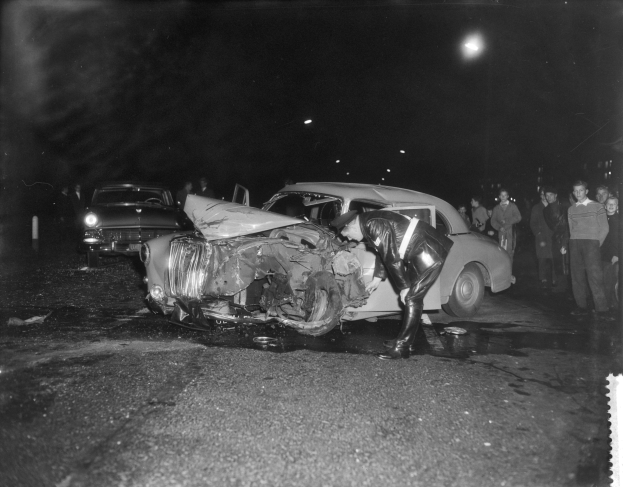 Schwarzes und weißes Foto eines beschädigten Autos am Straßenrand mit mehreren Menschen in der Nähe und beleuchteter Hintergrund.