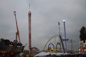 Eine Menschenmenge versammelt sich um einen hell erleuchteten Karneval in der Nacht, mit beleuchteten Fahrgeschäften, Pfählen und Bäumen unter einem bewölkten Himmel.