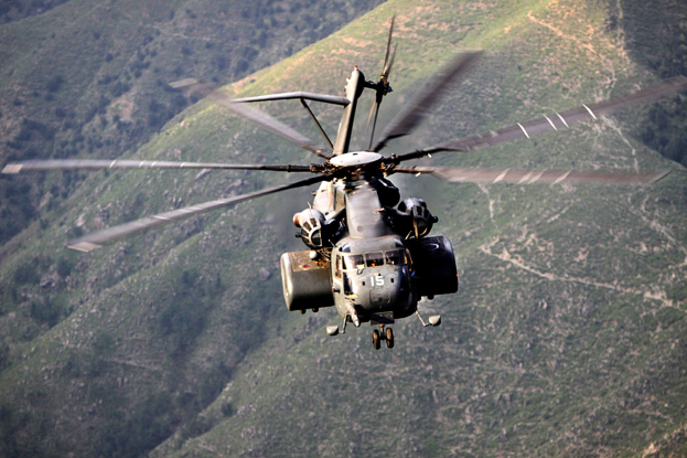 Ein Militärhelikopter mit ausgefahrenem Fahrwerk fliegt über ein Gebirgsmassiv mit grünen Hüllen im Hintergrund.