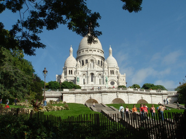 Ein weißes Gebäude umgeben von einem Zaun, Gras, Bäumen und Menschen, mit Pflanzen und Pfählen in der Nähe.