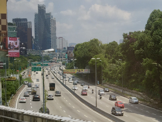 Eine belebte Autobahn mit hohen Gebäuden, Straßenlaternen, Leuchtschildern, Bäumen und einem bewölkten Himmel.