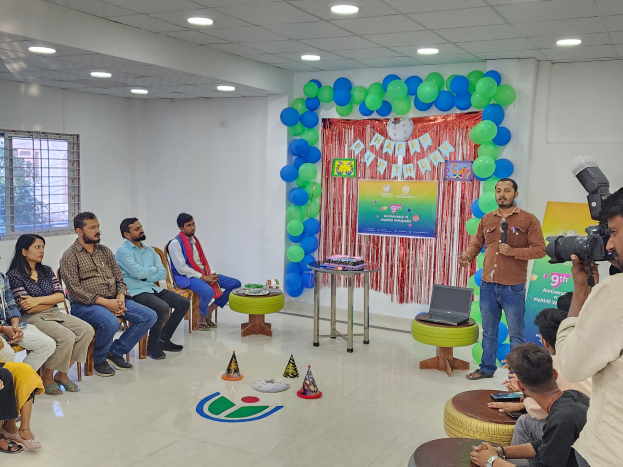 Eine Gruppe von Menschen, die auf Stühlen vor einem Publikum sitzen, mit einer Person, die ein Mikrofon hält, einem Laptop auf einem Hocker, einem Tisch mit verschiedenen Gegenständen, einem Banner, Luftballons, einem Vorhang, einem Fenster und einer Decke mit Deckenleuchten, die einen 9. Geburtstag feiern.
