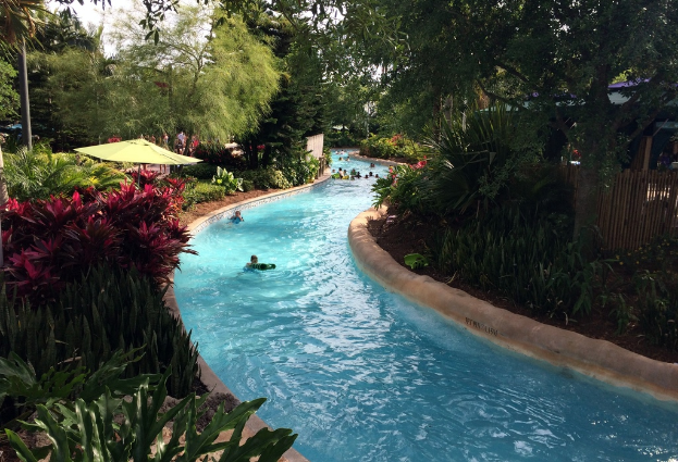 Ein Schwimmbad mit schwimmenden Menschen, umgeben von Pflanzen, Bäumen und einem Zaun, mit Menschen, Sonnenschirmen und Gebäuden im Hintergrund.