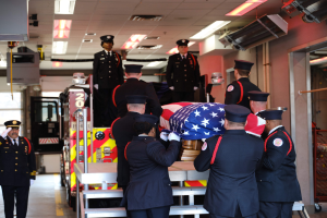 Eine Gruppe uniformierter Männer, die einen Sarg mit einer amerikanischen Flagge tragen, vor dem Hintergrund eines Feuerwehrautos und Deckenlampen.
