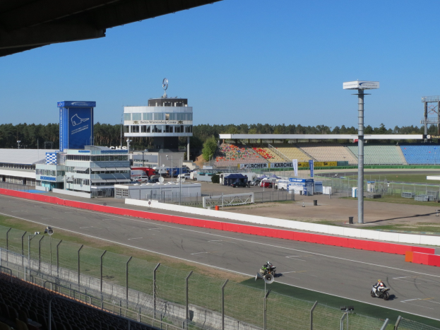 Eine Gruppe von Menschen auf Motorrädern auf einer Rennstrecke, umgeben von einem Zaun, Fahrzeugen, einem Gebäude, Schildern, Polen, einem Stadion, Lampen, Bäumen und einer bewölkten Himmel.
