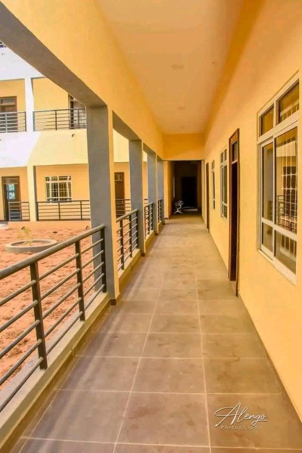 Ein Gebäude mit Fenstern, Türen, Säulen, Geländern und einem langen Flur mit einem gefliesten Boden und einem Balkon, mit anderen Gebäuden und einem klaren blauen Himmel im Hintergrund, und einem Text unten, der "3-Zimmer-Wohnung zur Miete in Ntinda, Kampala" liest.