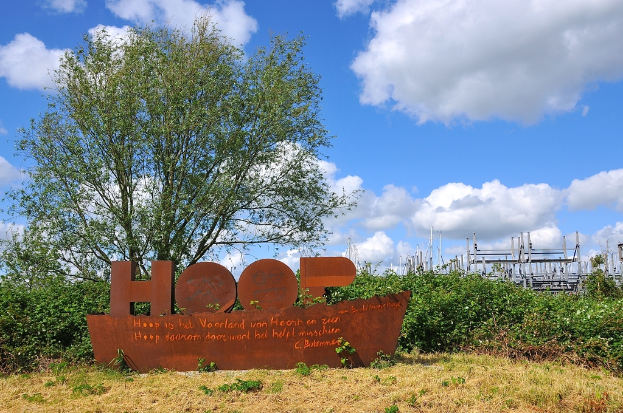 Ein Modellboot ist von Grün umgeben mit Pfählen im Hintergrund und trockenem Gras am Boden des Bildes.