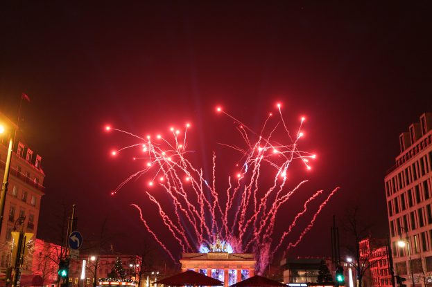 Eine Straßenansicht in Berlin am Silvesterabend, mit Gebäuden, Bäumen, Laternenmasten, Ampeln, Schildern, Zelten und Menschen, mit einem von Feuerwerk erleuchteten Himmel im Hintergrund.