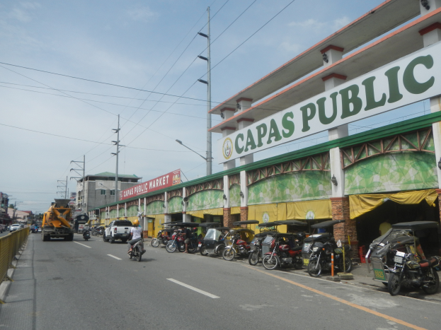 Eine belebte Stadtstraße mit Fahrzeugen, einem Geländer auf der linken Seite, Gebäuden, Pfählen, Laternen, Schildern und einigen Menschen unter einem bewölkten Himmel, mit einem Gebäude auf der Seite, das ein Schild trägt, auf dem "Capas Public" steht.