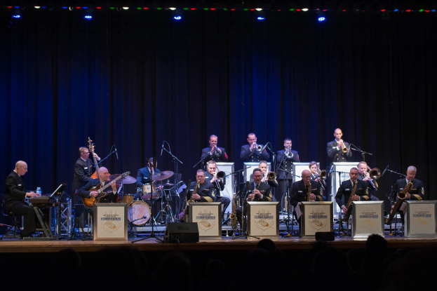 Eine Gruppe von Musikern auf einer Bühne, die Instrumente spielen, mit Mikrofonen und Ständern, Schilder mit der Aufschrift "The New Orleans Jazz Band", ein Vorhang im Hintergrund und ein Publikum darunter unter Bühnenlichtern.