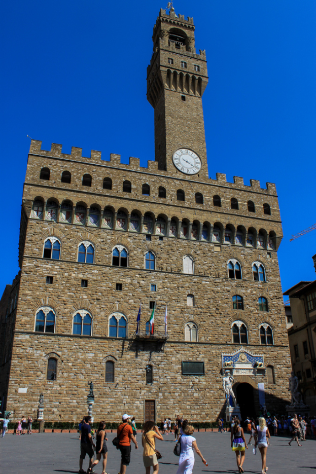 Personen im Vordergrund mit Gebäuden, Flaggen und einer Uhr an der Wand des zentralen Gebäudes im Hintergrund.