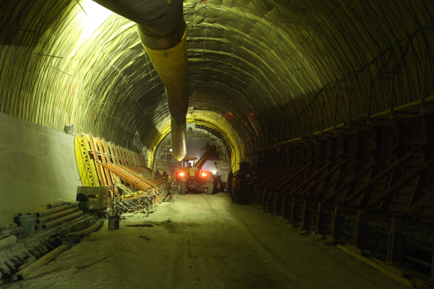 Eine Baustelle mit einem großen Tunnel in der Mitte, umgeben von Fahrzeugen, verstreuten Holzgegenständen, Rohren und einer Wand auf der linken Seite, mit einem Rohr oben und Hintergrundlichtern, die den Tunnel beleuchten.