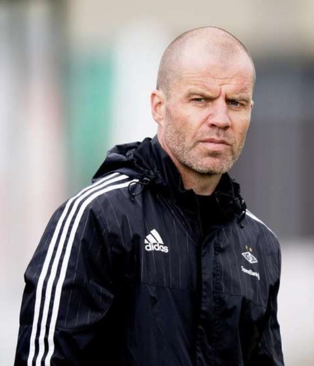 Ein kahlgeschorener Trainer in einer blauen Jacke mit weißen Streifen und Kragen steht auf einem Fussballfeld und schaut ernst in die Ferne.