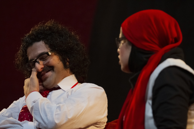 Zwei Personen, ein Mann in einem weißen Hemd mit roter Krawatte und eine Frau mit Brille, sitzen nebeneinander auf einer dunklen Bühne.