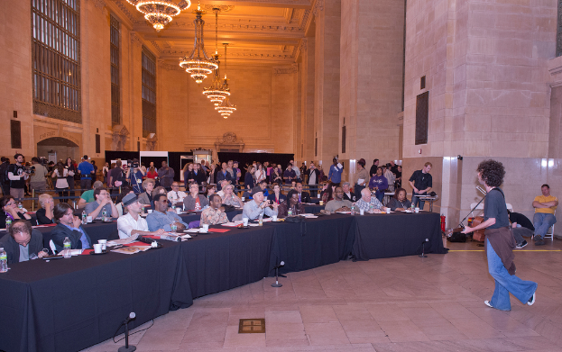 Eine Konzertszene mit einem Gitarristen und Sänger auf der rechten Seite, einem beleuchteten Bühnenbereich und einem Publikum im Hintergrund.