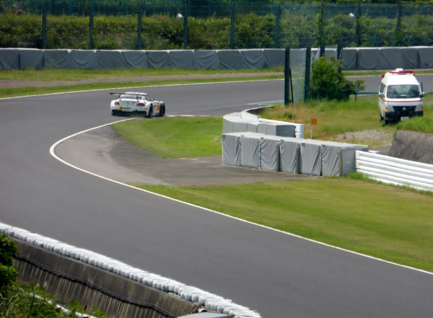 Ein Rennauto fährt eine Rennstrecke entlang, im Hintergrund ist ein Polizeiwagen zu sehen, umgeben von Gras, Pflanzen, Bäumen und einer Absperrung, rechts steht ein weiteres Fahrzeug, unten steht Text.