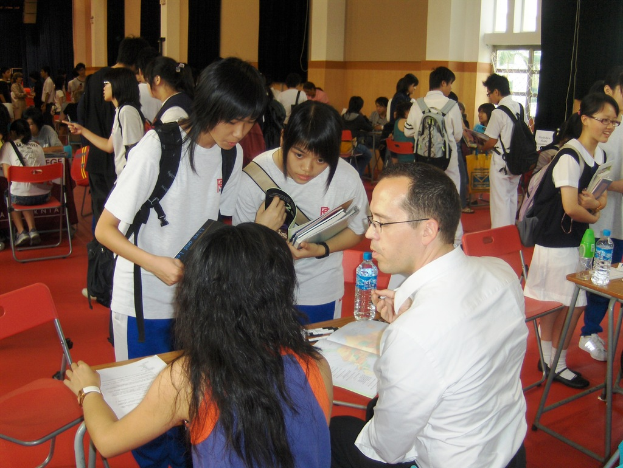Ein Klassenzimmer mit mehreren Mädchen in weißen T-Shirts, die Bücher in der Hand halten, vorne, ein Mann und eine Frau, die unten sitzen und diskutieren, und eine gelbe Wand mit einem Glasfenster im Hintergrund.