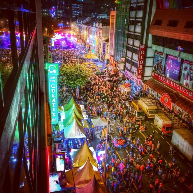 Eine belebte Jahrmarktszene mit einer großen Menschenmenge auf der Straße, umgeben von Zelten, Geschäften, Gebäuden und Bannern, mit zahlreichen Lichtern im Hintergrund.