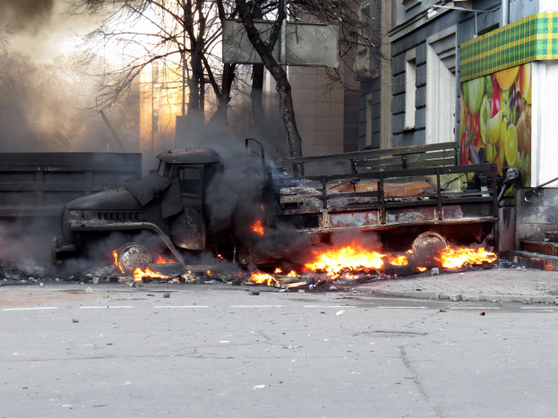 Ein auf der Straße stehender, brennender Lkw mit qualmenden Rauchwolken, Bäume, Gebäude und einen klaren Himmel im Hintergrund.