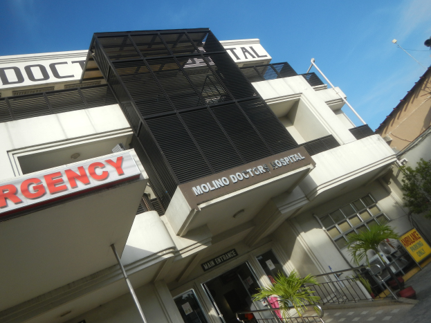 Ein Gebäude mit einer "Notfall"-Aufschrift an der Seite, umgeben von Pflanzen, einem Metallzaun, Bäumen und Pfählen, unter einem bewölkten Himmel.