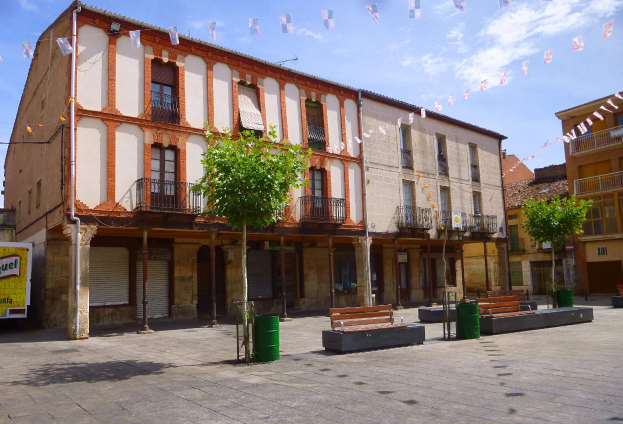 Ein Plaza Mayor mit mehreren Gebäuden mit Fenstern, Geländern, Bäumen, Bänken, Mülltonnen, Pfählen, Drähten und dekorativen Flaggen in der Mitte, unter einem klaren blauen Himmel.